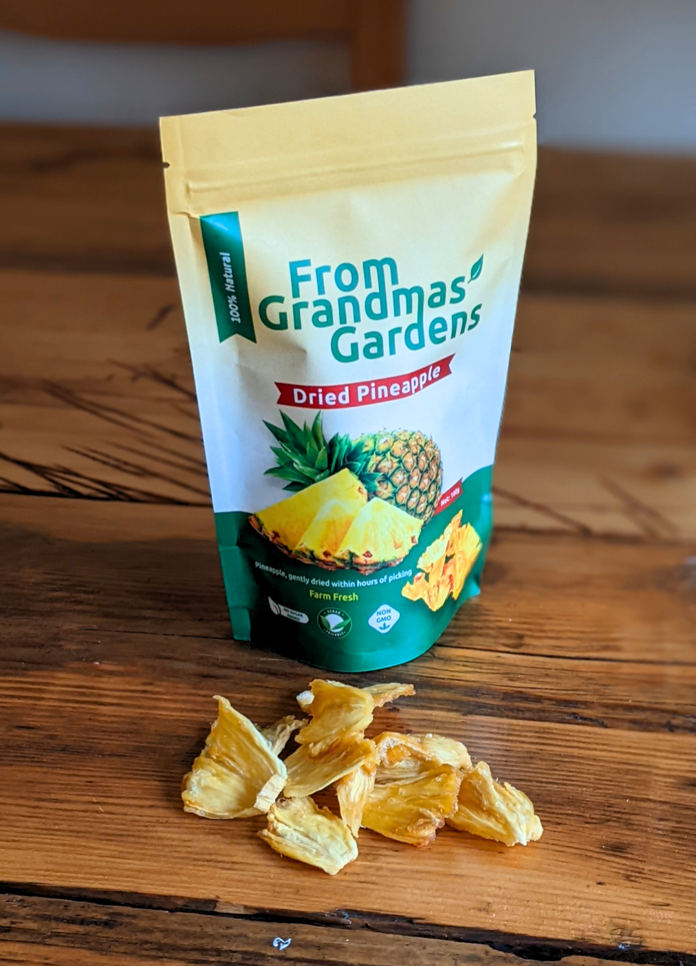 100g pouch of From Grandmas' Gardens Dried Pineapple with a small pile of dried pineapple slices on the wooden table in front.